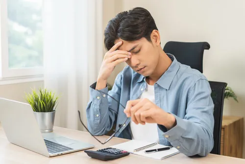 exhausted young businessman holding glasses at desk with laptop and calculator